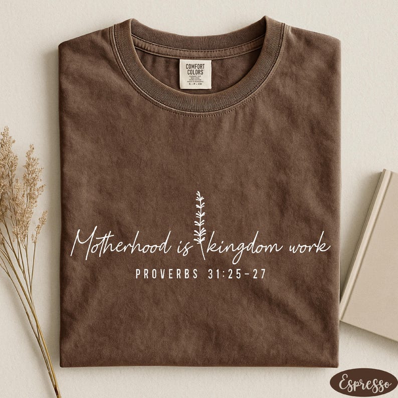 A brown t-shirt with a printed message about motherhood and kingdom work, along with a small plant and a book.