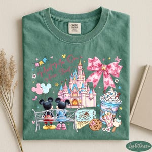 A green t-shirt with a Disney-themed design, including Mickey Mouse, a castle, and various Disney-related items such as a cup of ice cream and a popcorn bucket.
