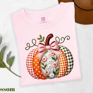 A pink t-shirt with a pumpkin embroidered on it, featuring a bow and flowers, is displayed alongside a brown hat and some greenery.