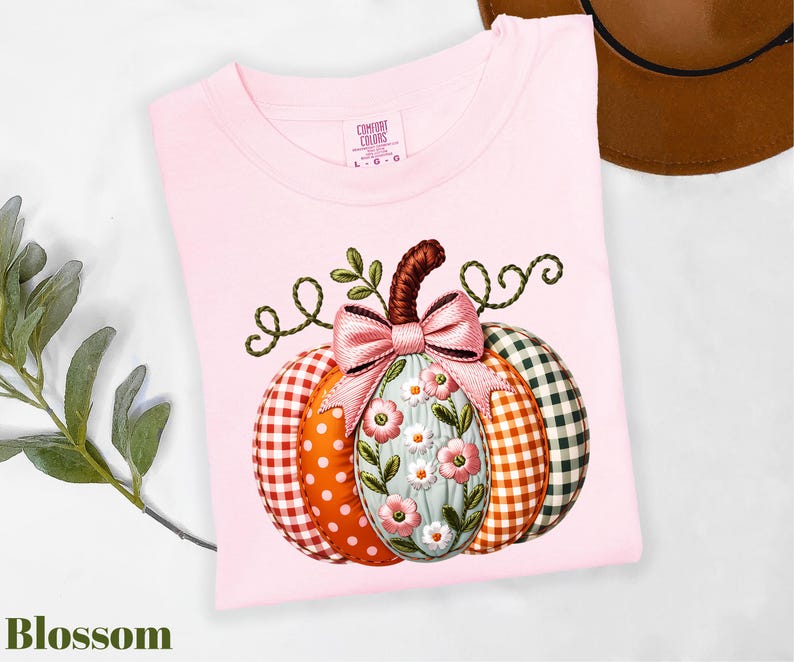 A pink t-shirt with a pumpkin embroidered on it, featuring a bow and flowers, is displayed alongside a brown hat and some greenery.