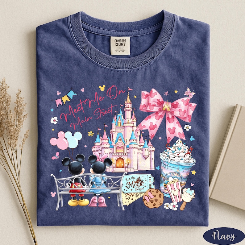 A navy blue t-shirt with a colorful graphic design depicting Mickey Mouse and Minnie Mouse sitting on a bench, surrounded by various Disney-themed elements such as a castle, popcorn, and ice cream.