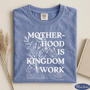 A blue t-shirt with white text and floral designs, along with a book and some dried flowers, arranged on a white surface.