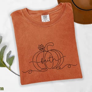 a red t-shirt with a pumpkin and the word "faith" printed on it, along with a brown hat and some greenery.
