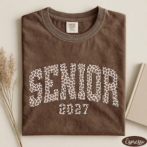 May include: A brown t-shirt with the word "SENIOR" in large, leopard print letters and the year "2027" underneath. The shirt has a crew neck and a tag that says "Comfort Colors". The shirt is folded on a light surface.