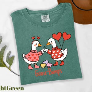 A green t-shirt with a cartoon illustration of two cartoon geese wearing heart-shaped sweaters and holding heart-shaped balloons, with the text "Goosebumps" below the image.