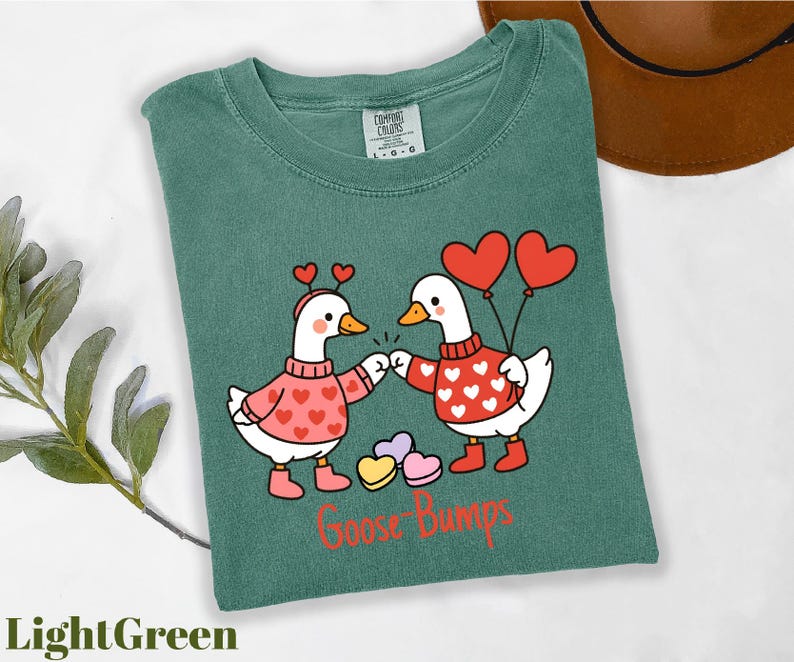 A green t-shirt with a cartoon illustration of two cartoon geese wearing heart-shaped sweaters and holding heart-shaped balloons, with the text "Goosebumps" below the image.