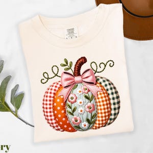 A white t-shirt with a colorful pumpkin embroidered on it, featuring a bow and floral elements. The t-shirt is placed on a white surface, with a brown hat and some greenery visible in the background.