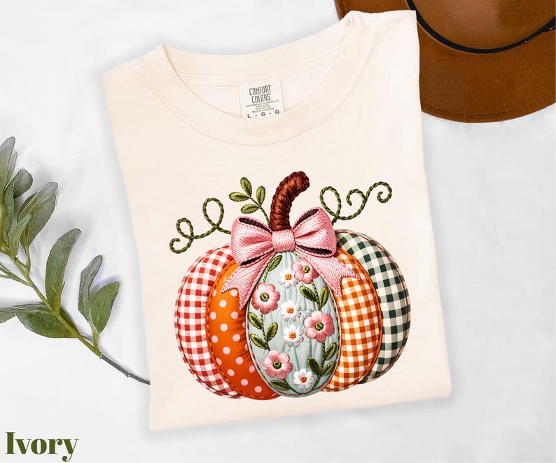 A white t-shirt with a colorful pumpkin embroidered on it, featuring a bow and floral elements. The t-shirt is placed on a white surface, with a brown hat and some greenery visible in the background.