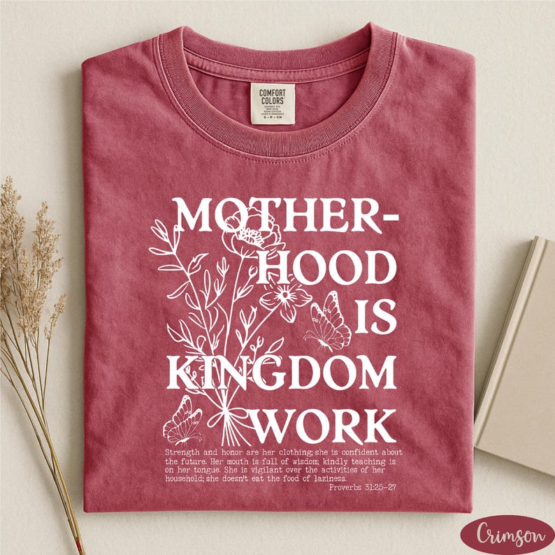 A red t-shirt with white text and floral designs, along with a book and some dried flowers, arranged on a white surface.