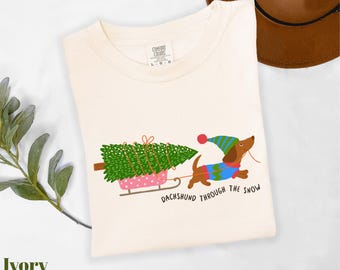 May include: Ivory-colored t-shirt featuring a dachshund pulling a Christmas tree on a pink sled. The dog wears a blue and green sweater and hat. Text reads "Dachshund Through The Snow".
