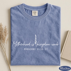 A blue t-shirt with white text and a small plant design, placed on a beige surface alongside a book and some dried flowers.