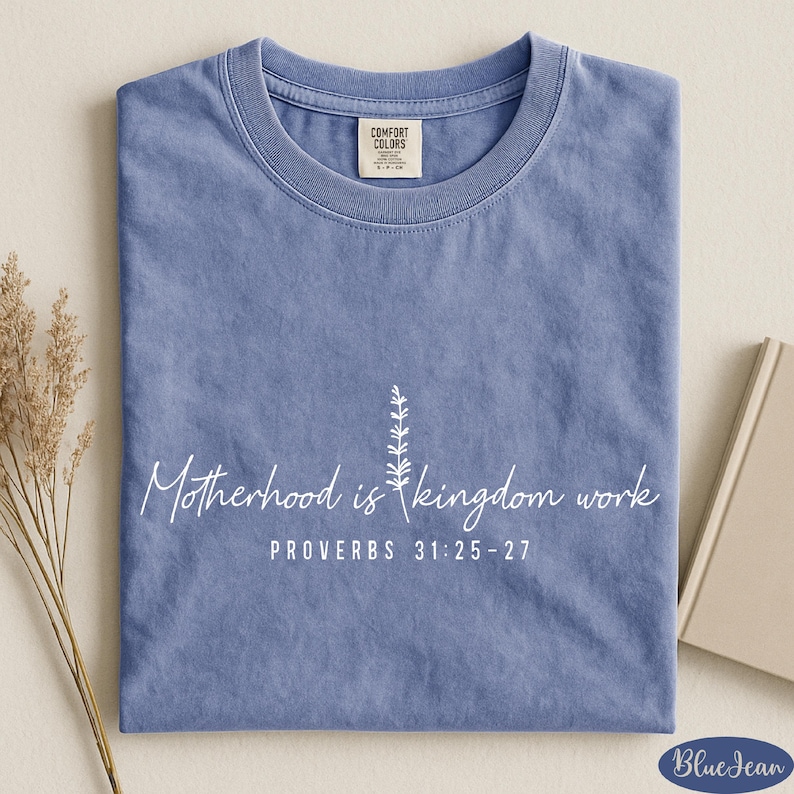 A blue t-shirt with white text and a small plant design, placed on a beige surface alongside a book and some dried flowers.