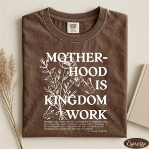 A brown t-shirt with a white graphic design that reads "MOTHER-HOOD IS KINGDOM WORK" and includes a Bible verse from Proverbs 3:25-29.