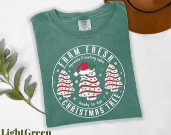 May include: Light green t-shirt with a circular graphic that reads "FARM FRESH CHRISTMAS TREE" with illustrations of Christmas trees and a Santa hat. The text "Sprinkle Frosting cake" and "Ready to Eat" are also included in the design.