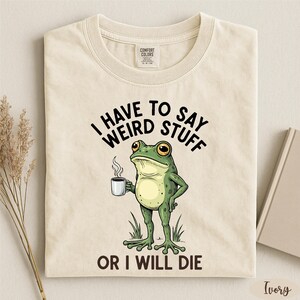 a white t-shirt with a cartoon frog holding a cup of coffee and the text "I HAVE TO SAY WEIRD STUFF OR I WILL DIE" printed on it.