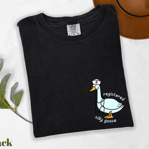 A black t-shirt with a cartoon image of a white goose wearing a nurse's hat and the text "registered silly goose" printed on it, along with a brown hat and some greenery.