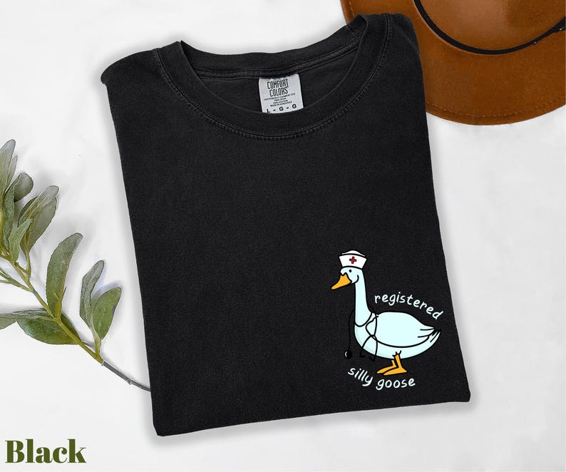 A black t-shirt with a cartoon image of a white goose wearing a nurse's hat and the text "registered silly goose" printed on it, along with a brown hat and some greenery.