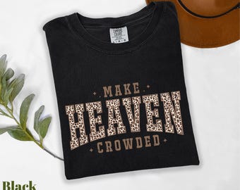 May include: Black t-shirt with the text "MAKE HEAVEN CROWDED" in a brown, leopard print design. The shirt is folded and has a tag in the upper center. A brown hat is visible in the background.