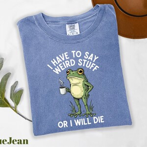 A blue t-shirt with a cartoon frog holding a coffee cup and the text "I HAVE TO SAY WEIRD STUFF OR I WILL DIE" printed on it.