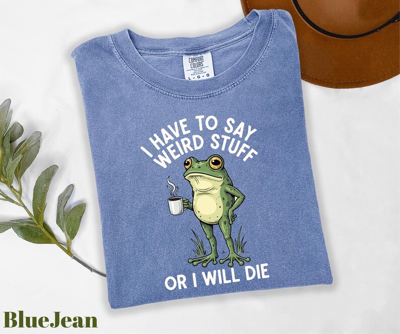 A blue t-shirt with a cartoon frog holding a coffee cup and the text "I HAVE TO SAY WEIRD STUFF OR I WILL DIE" printed on it.
