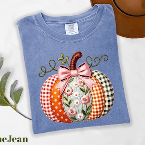 A blue denim t-shirt with a colorful pumpkin embroidered on it, along with a brown hat and some greenery, is displayed on a white surface.