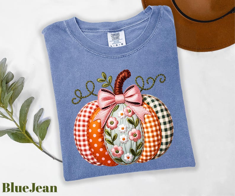 A blue denim t-shirt with a colorful pumpkin embroidered on it, along with a brown hat and some greenery, is displayed on a white surface.