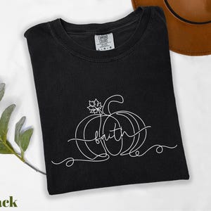 A black t-shirt with a white pumpkin design and the word "Faith" written on it, along with a brown hat and some greenery, all placed on a white surface.