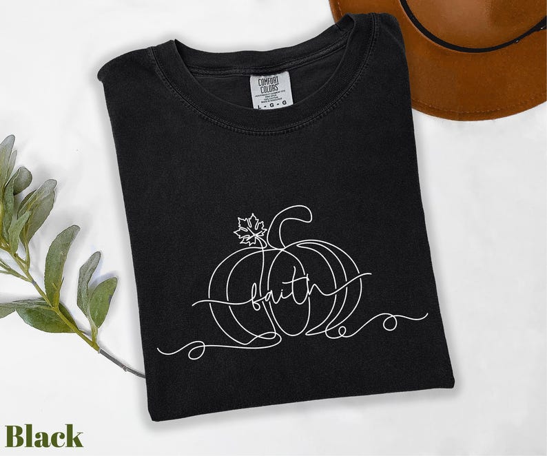 A black t-shirt with a white pumpkin design and the word "Faith" written on it, along with a brown hat and some greenery, all placed on a white surface.