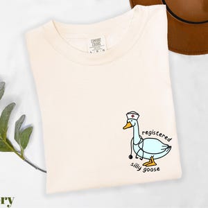 A white t-shirt with a cartoon image of a duck wearing a nurse's hat and the text "registered silly goose" printed on it, along with a brown hat and some greenery.