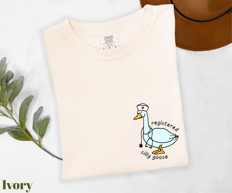 A white t-shirt with a cartoon image of a duck wearing a nurse's hat and the text "registered silly goose" printed on it, along with a brown hat and some greenery.