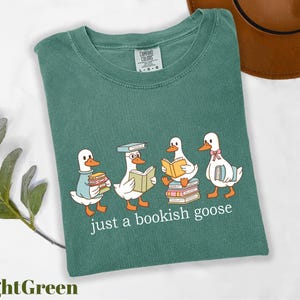 May include: A light green t-shirt featuring a graphic of four cartoon geese reading books. The text "just a bookish goose" is printed below the geese. The shirt is made of a soft material.