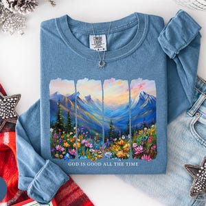 May include: A blue long-sleeve shirt with a mountain and floral landscape design. The graphic features a colorful mountain range, trees, and flowers, with the text "GOD IS GOOD ALL THE TIME" at the bottom. A silver necklace is visible.