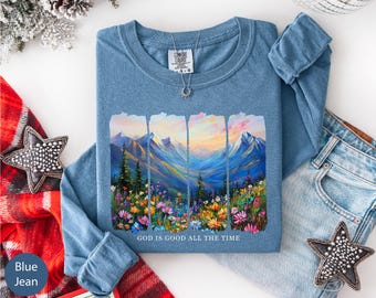 May include: A blue long-sleeve shirt with a mountain and floral landscape design. The graphic features a colorful mountain range, trees, and flowers, with the text "GOD IS GOOD ALL THE TIME" at the bottom. A silver necklace is visible.