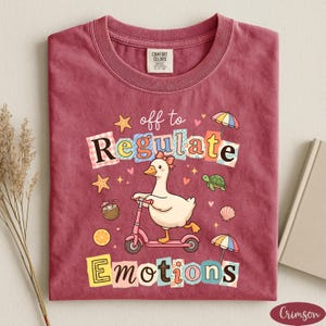 a red t-shirt with a cartoon duck riding a pink scooter, surrounded by various colorful objects such as umbrellas, flowers, and a book.