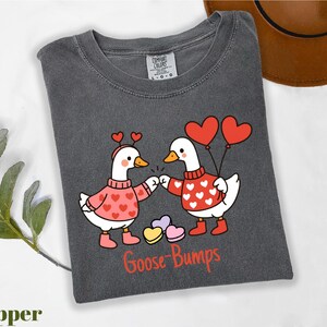 A gray t-shirt with a graphic design depicting two cartoon geese wearing heart-shaped sweaters and holding heart-shaped balloons, with the text "Goose Bumps" below the image.