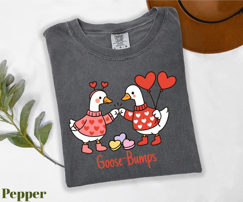 A gray t-shirt with a graphic design depicting two cartoon geese wearing heart-shaped sweaters and holding heart-shaped balloons, with the text "Goose Bumps" below the image.