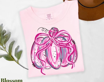 Può includere: Una t-shirt rosa chiaro con la grafica di una zucca decorata con un nastro rosa brillante. La zucca presenta texture a pennellate nei toni del rosa, bianco e grigio con contorni neri. La t-shirt ha un'etichetta Comfort Colors sul collo.