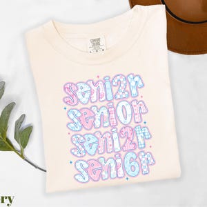 A white t-shirt with a colorful text design that says "senior senior senior" printed on it, along with some decorative elements.