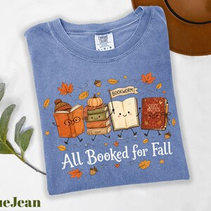 A blue t-shirt with a graphic design of four anthropomorphic books, each with a unique character and fall-themed elements, such as leaves and a pumpkin. The text on the t-shirt reads "All Booked for Fall".