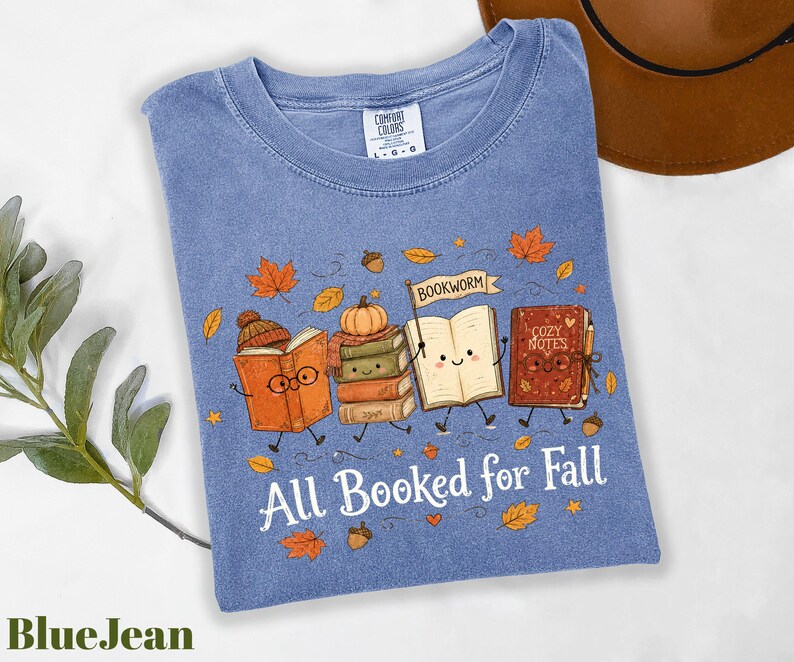 A blue t-shirt with a graphic design of four anthropomorphic books, each with a unique character and fall-themed elements, such as leaves and a pumpkin. The text on the t-shirt reads "All Booked for Fall".