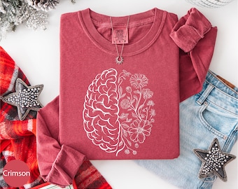 May include: Crimson long-sleeve shirt with a white graphic of a brain and flowers. The shirt has a crew neck and a small tag. A silver necklace with a sun pendant is visible. The shirt is folded next to a pair of blue jeans.