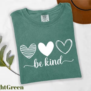 A green t-shirt with the text "be kind" printed on it, along with three heart symbols, is displayed on a white background.
