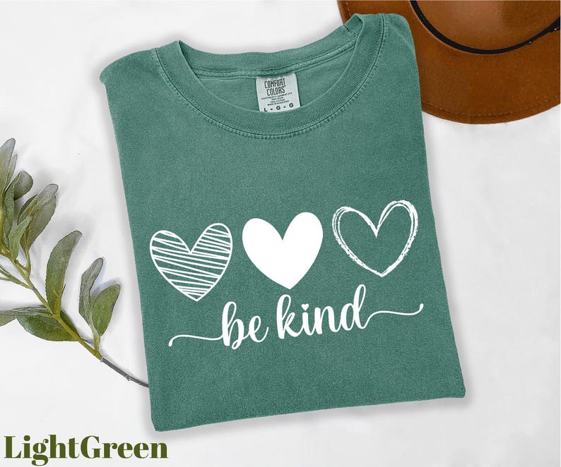 A green t-shirt with the text "be kind" printed on it, along with three heart symbols, is displayed on a white background.