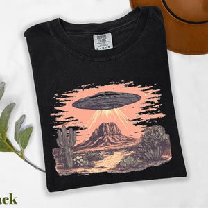 a black shirt with a picture of an alien flying over a desert