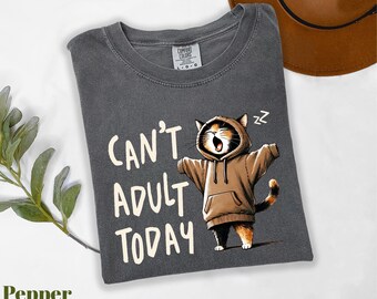 May include: A dark gray t-shirt with the text "Can't Adult Today" and a cartoon cat wearing a brown hoodie. The cat is yawning with its arms outstretched. A brown hat is in the background.