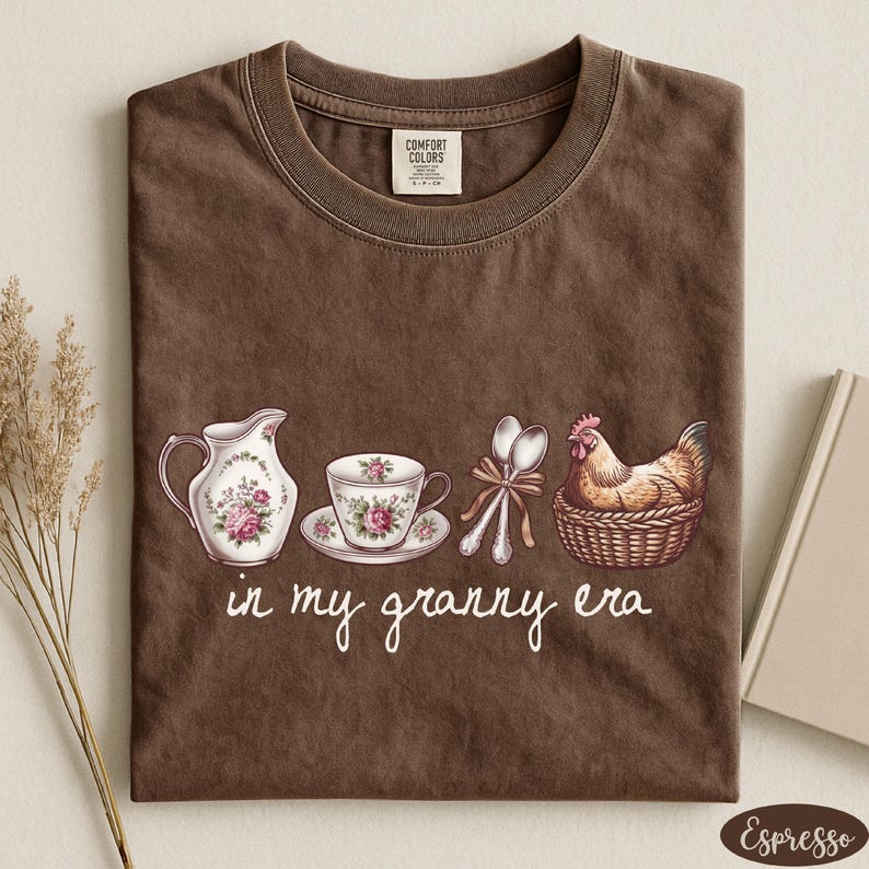 A brown t-shirt with a printed design of a tea set and a chicken in a basket, along with the text "in my granny era" printed on it.