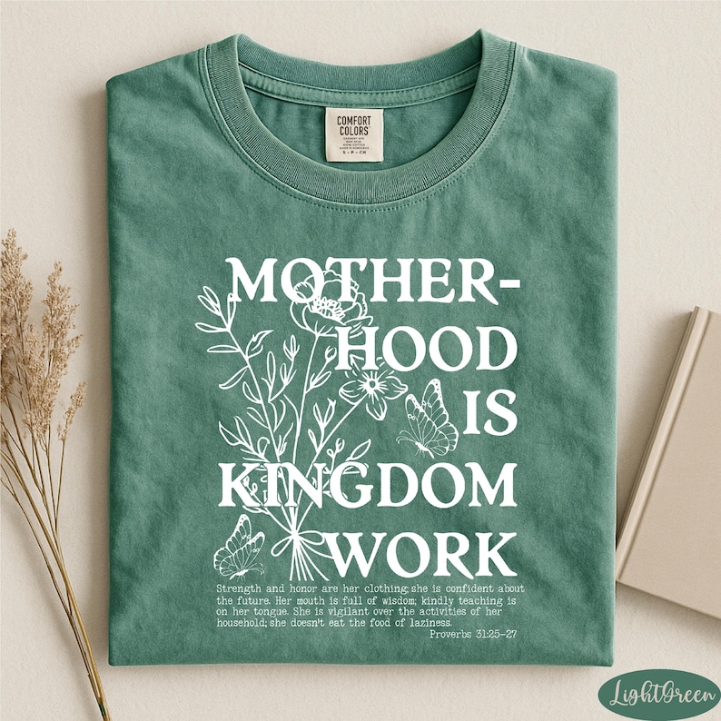 A green t-shirt with white text and floral designs, along with a book and some dried flowers, arranged on a white surface.