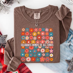 a brown t-shirt with a colorful floral design and the text "Celebrate Minds of All Kinds" printed on it, along with a necklace and a pair of jeans.