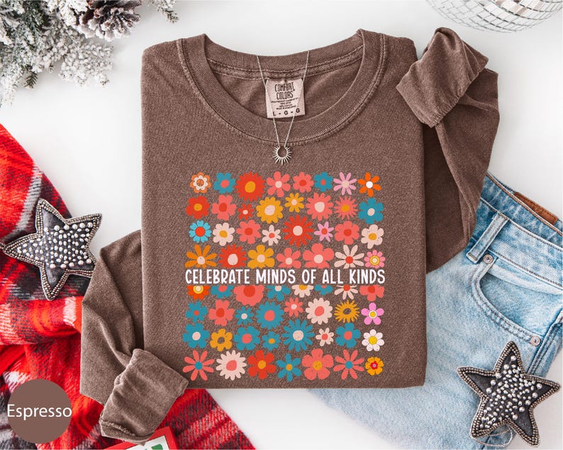 a brown t-shirt with a colorful floral design and the text "Celebrate Minds of All Kinds" printed on it, along with a necklace and a pair of jeans.