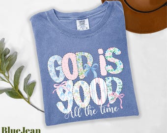 May include: Blue jean colored t-shirt with the words "GOD IS GOOD" in a floral and pastel design. The phrase "All the time" is written in a cursive font below the main text. A brown hat is visible in the background.